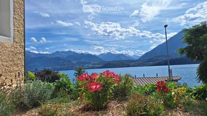 Garden with red flowers, shrubs, and a view of a lake and mountains in the background.