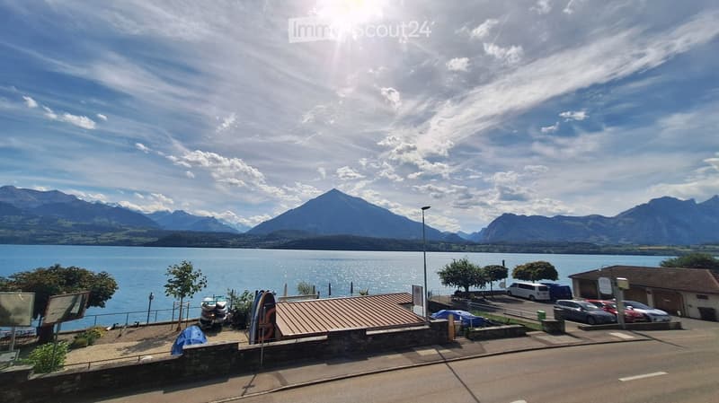 View of a lake with mountains in the background, parking area with cars, and a small building near the water.