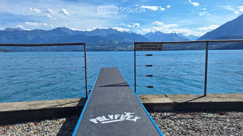 A view of a lake with a metal railing and a sign in German, with mountains in the background.