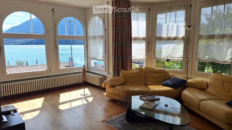 Bright living room with large windows overlooking a lake and mountains, featuring a cream-colored sofa, glass coffee table, and floor lamp.