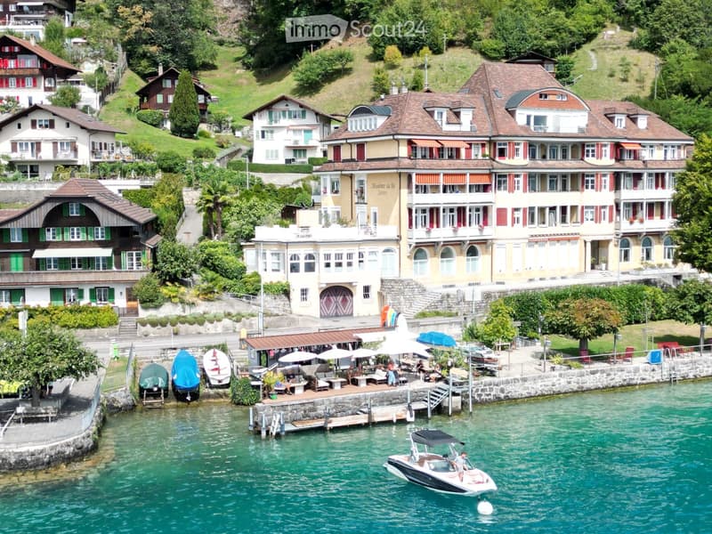 A scenic lakeside village with multiple multi-story houses, balconies, and a waterfront promenade with boats and outdoor seating.