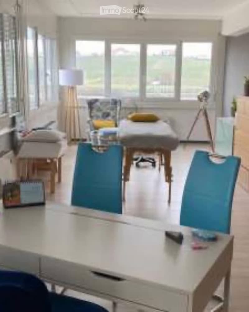Brightly lit room with large windows, a massage table, chairs, a sofa, and a lamp. The space is well-organized and appears to be used for relaxation or therapy.