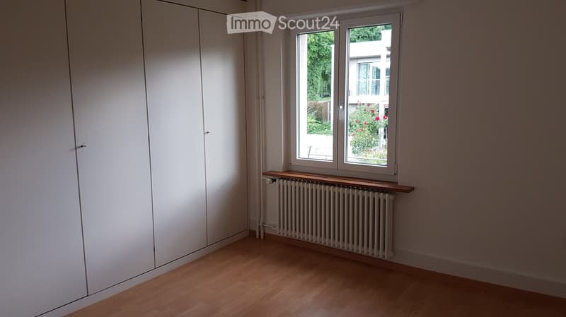 Empty room with wooden flooring, white walls, large window with view of greenery, built-in wardrobe, radiator, and wooden shelf.