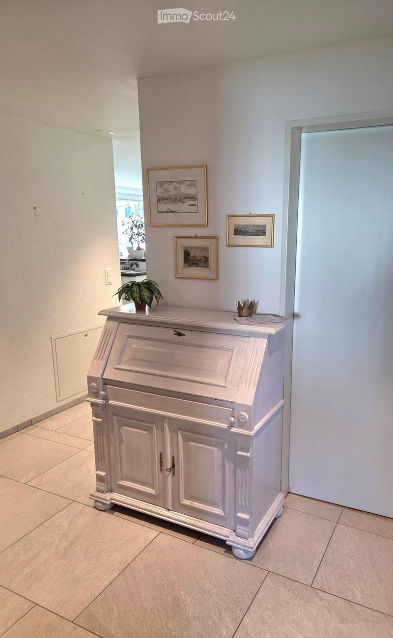 A hallway with a white console table, framed artwork, and a small potted plant. The floor is tiled, and the walls are painted white.