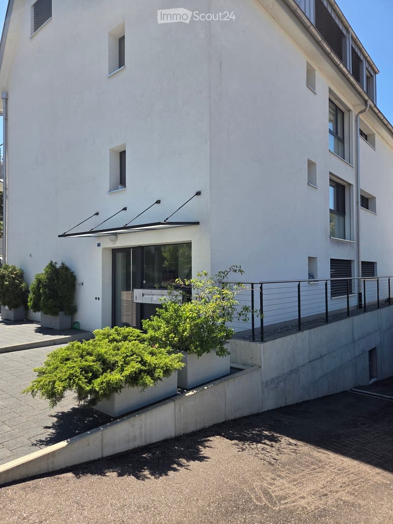 Modern two-story building with white facade, multiple windows, and a covered entrance area with a railing and potted plants.