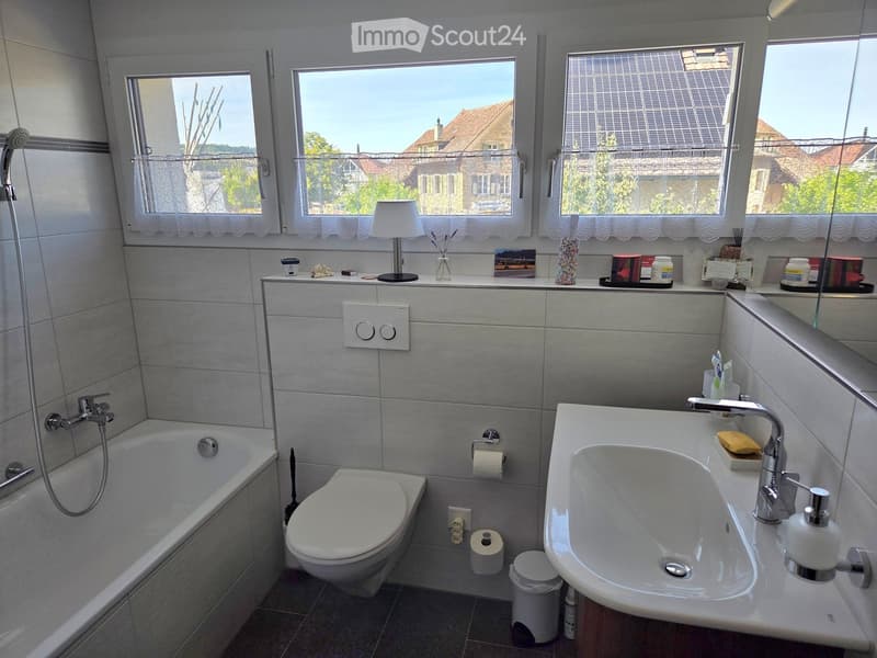 Modern bathroom with a bathtub, toilet, sink, and showerhead. Features a large window with white curtains, a small table lamp, and various toiletries on the windowsill.