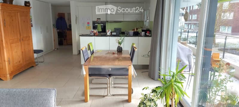 Bright, modern dining area with a wooden dining table, chairs, and a view of a balcony. The space includes a kitchen area with cabinets and an oven, and a large wooden cupboard is visible on the left side.