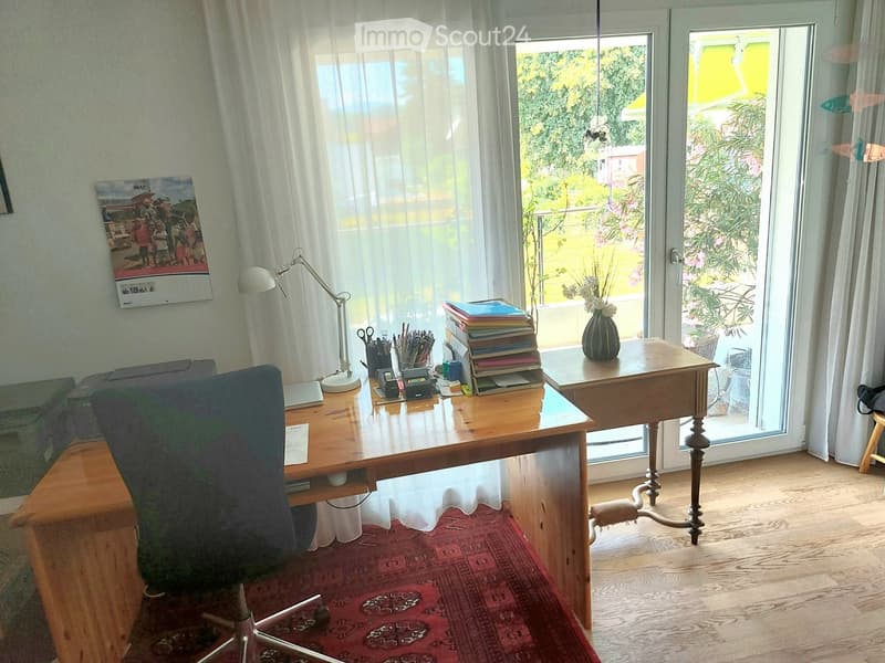 Brightly lit room with wooden floors, large windows, and a desk with a chair, lamp, and various office supplies.