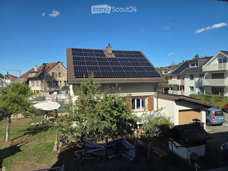 A two-story house with solar panels on the roof, surrounded by trees and a small garden area with a table and umbrella.