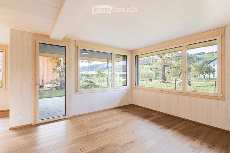 Bright, empty room with wooden flooring and light wooden walls, featuring large windows and a sliding glass door leading to an outdoor area.