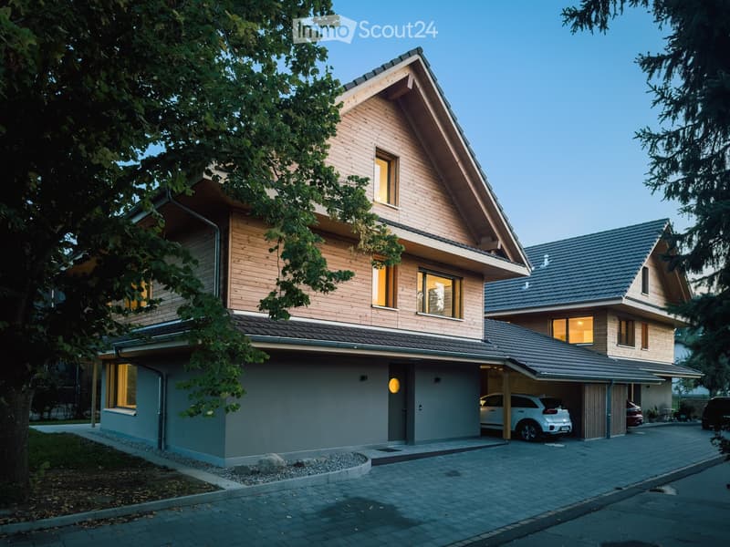 Two-story modern house with wooden facade, grey garage, and multiple windows. Features a driveway with parked cars and surrounding greenery.