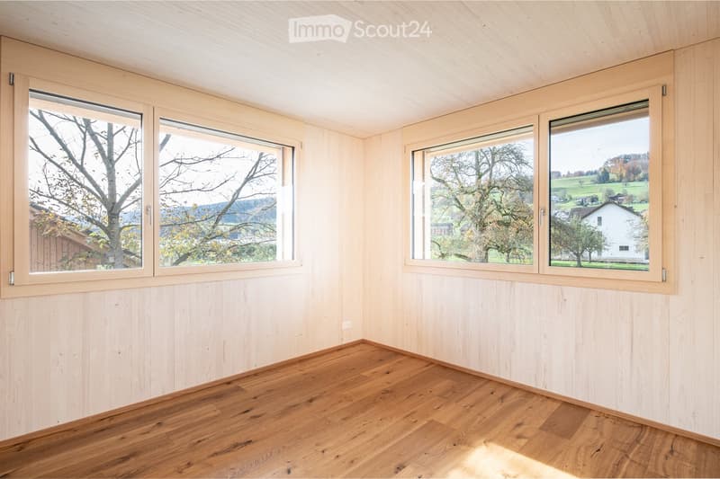 Empty room with light wooden walls, wooden floor, and large windows offering a scenic view of trees and countryside.