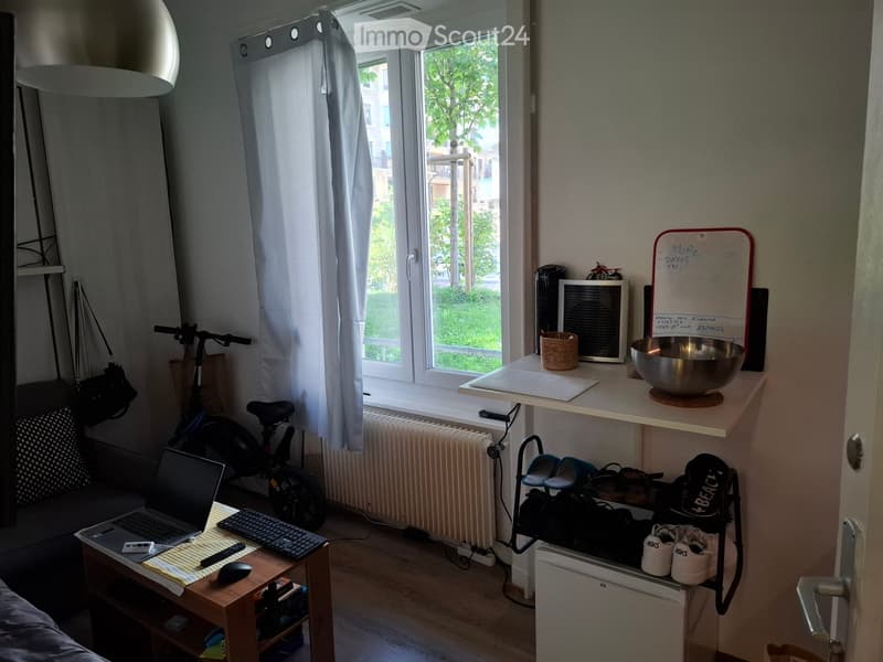 A small, well-lit room with a wooden floor, a sofa, a laptop on a small table, a bicycle, a scooter, a refrigerator, and a shelf with various items including a bowl and a board.