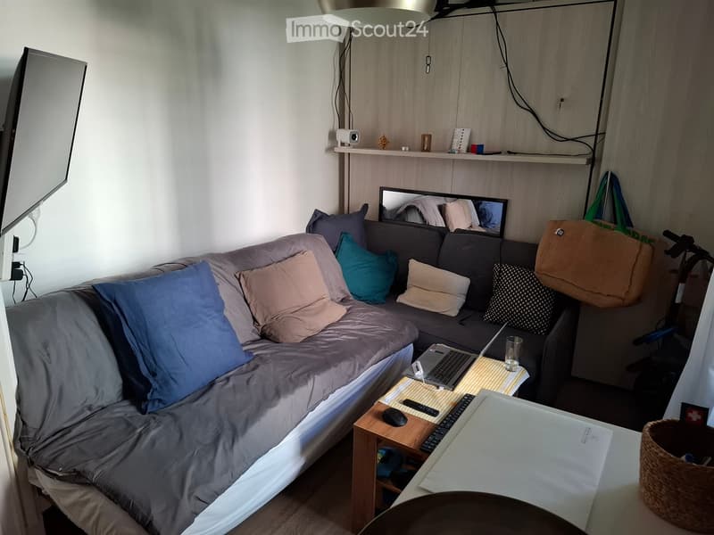 A cozy living space with a sofa, laptop on a small table, and a mirror above the sofa. The room features a modern design with a mix of wooden and white elements.