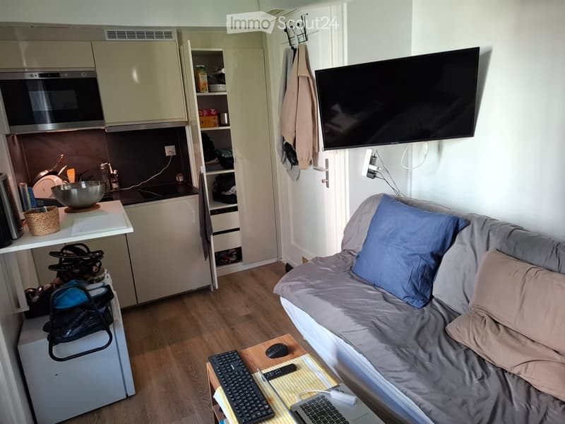 A compact studio apartment with a sofa, a small kitchen area, a TV mounted on the wall, and a desk with a laptop and other items.
