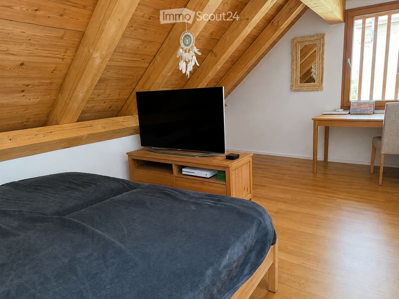 Cozy attic bedroom with wooden slanted ceilings, a large flat-screen TV on a wooden stand, a neatly made bed with dark blue bedding, a small desk with a chair, and a decorative dreamcatcher hanging from the ceiling.