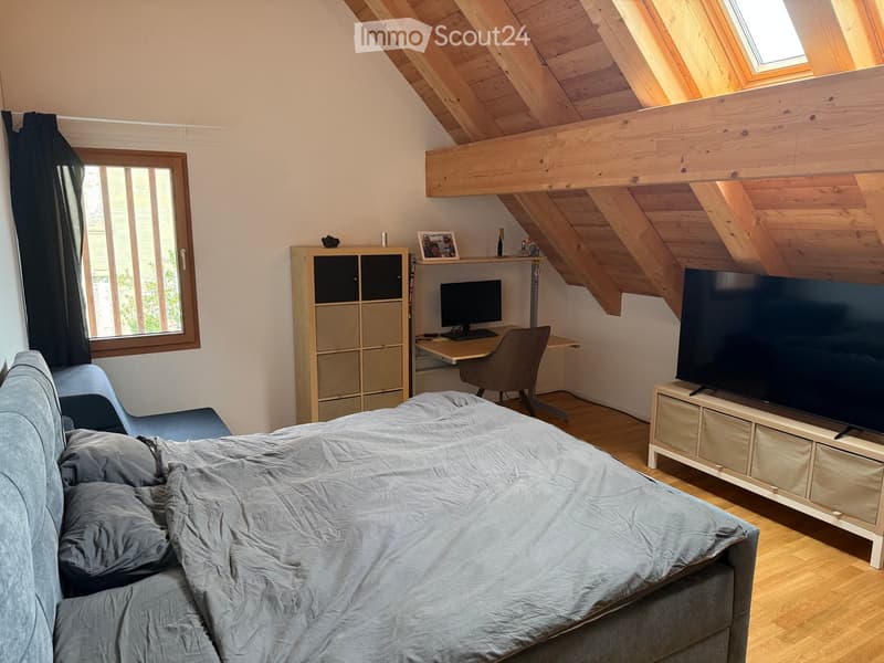 Cozy attic bedroom with wooden slanted ceilings, a large window, a bed with grey bedding, a sofa, a TV, a desk with a computer, and a cabinet.