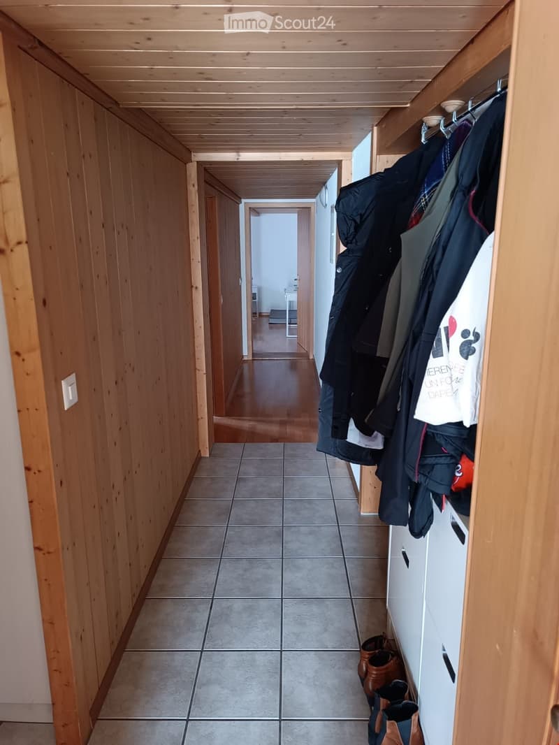 A narrow hallway with wooden paneling and a tiled floor, featuring a coat rack with jackets and shoes, and a visible room at the end.