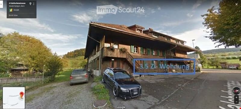A large, two-story wooden house with a brown roof and multiple windows, situated in a rural area with a parking area in front.