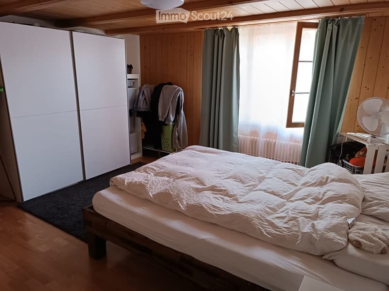 Cozy bedroom with wooden walls and ceiling, featuring a large bed with white bedding, a wardrobe, a fan, and a window with curtains.
