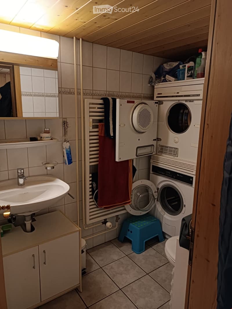 Compact bathroom with a sink, mirror, towel rack, washing machine, and toilet. Wooden ceiling and tiled walls.