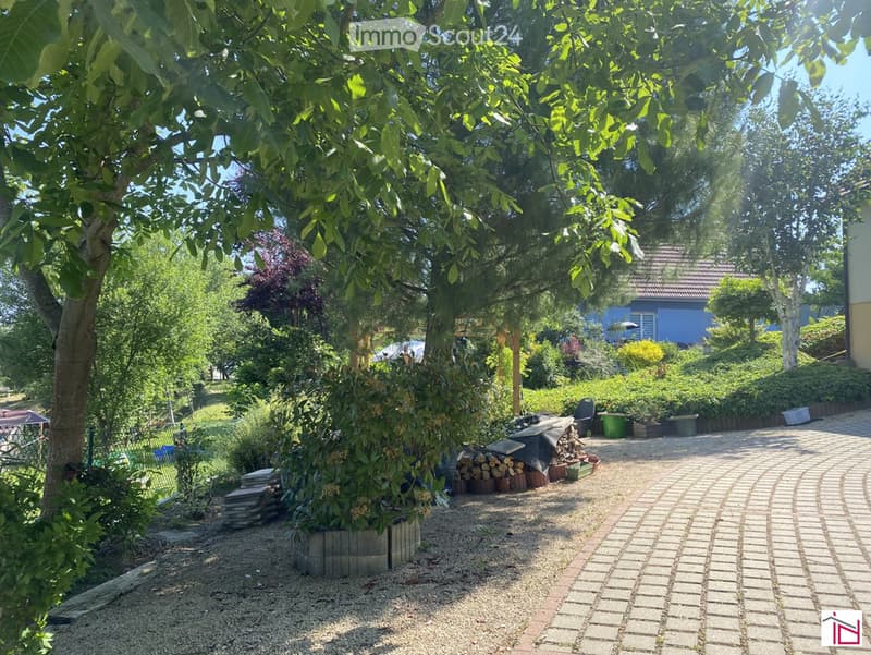 Outdoor area with trees, shrubs, and garden beds, gravel pathway, and a house in the background.