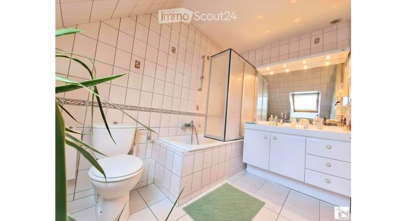 Modern bathroom with white tiles, a bathtub, toilet, sink, and a large mirror with built-in lighting. Features a glass-enclosed shower and a green bath mat on the floor.