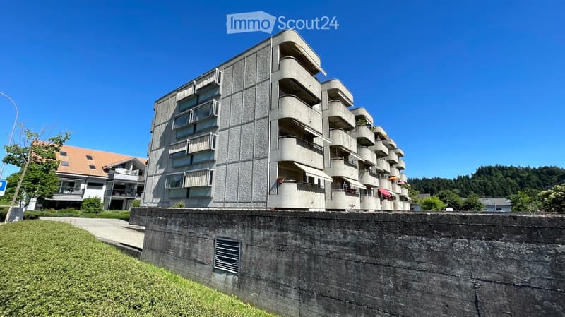 Modern multi-story apartment building with balconies and large windows, surrounded by greenery and a concrete retaining wall.