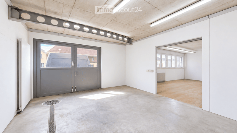 Empty garage with concrete floor, white walls, large grey garage door, and a view of a van outside. The adjacent room has wooden flooring and white walls.