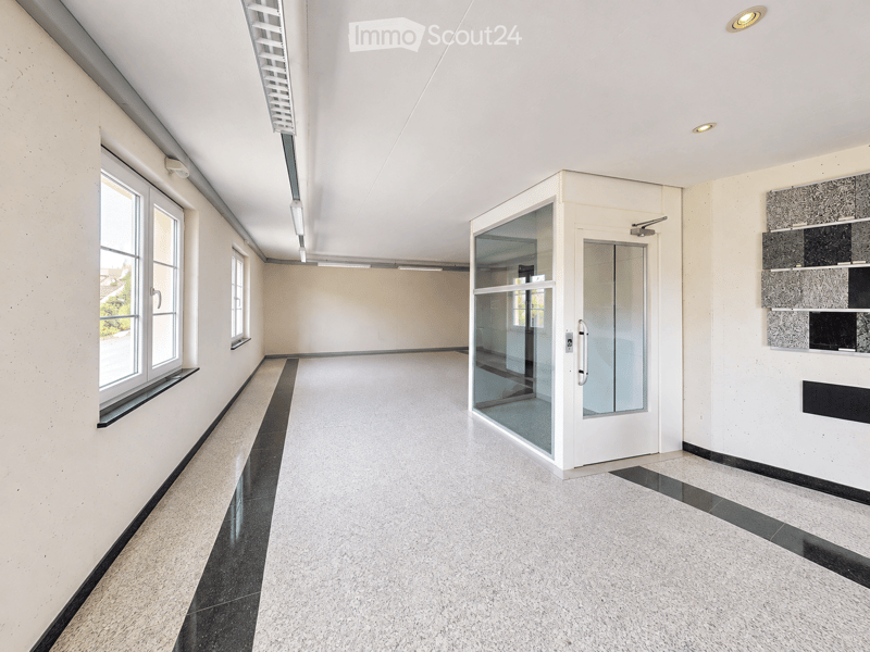 Empty hallway with white walls, large windows, glass door, and modern lighting fixtures.