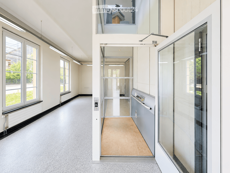 Modern, clean hallway with large windows, glass doors, and a raised platform leading to an upper level. The space features a smooth, light-colored floor and white walls.