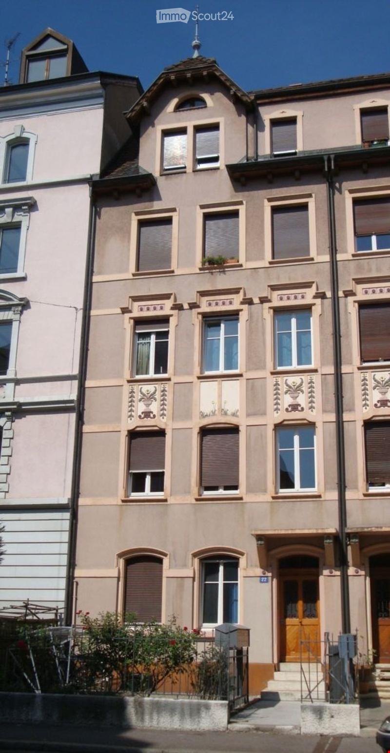 A multi-story residential building with beige and light pink facade, multiple windows with shutters, decorative moldings, and a small garden area in front.
