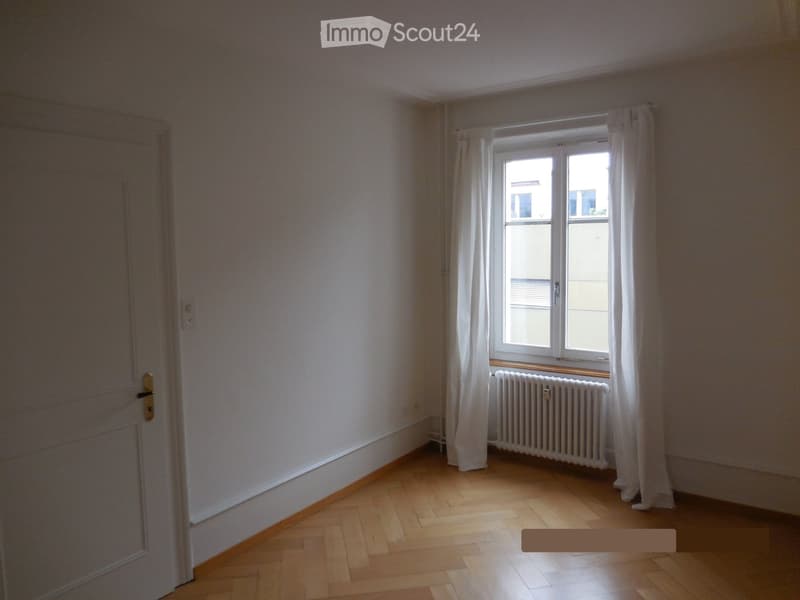 Empty room with wooden flooring, white walls, a window with white curtains, and a radiator below the window.