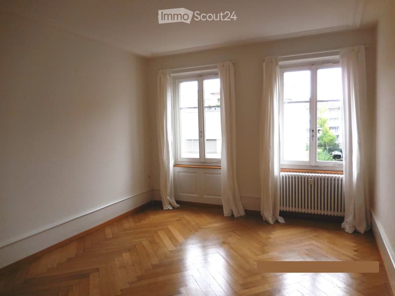 Empty room with wooden flooring, large windows with white curtains, and radiators beneath the windows.