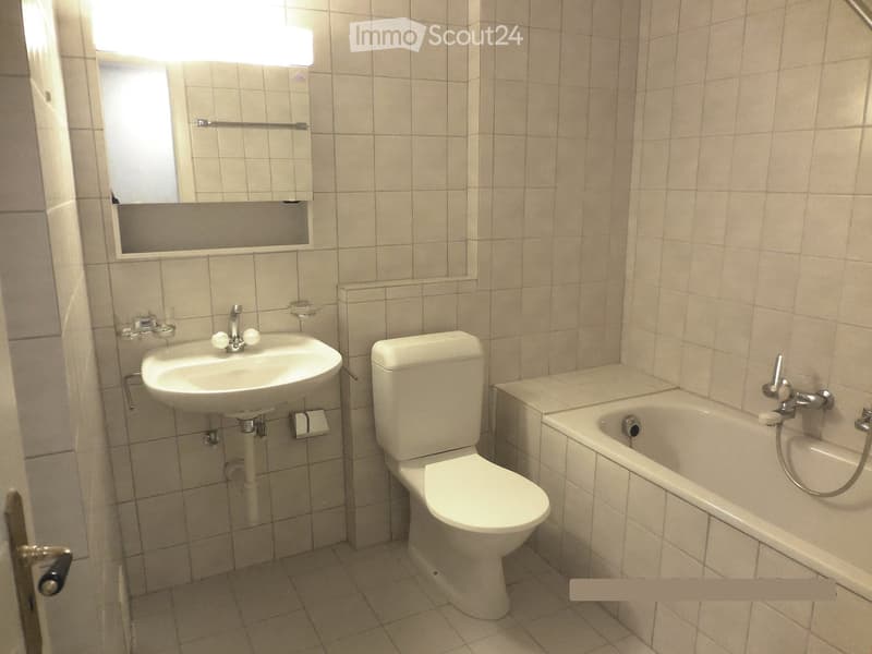 A small bathroom with beige tiled walls and floor, featuring a sink, toilet, bathtub, and a mirror above the sink.