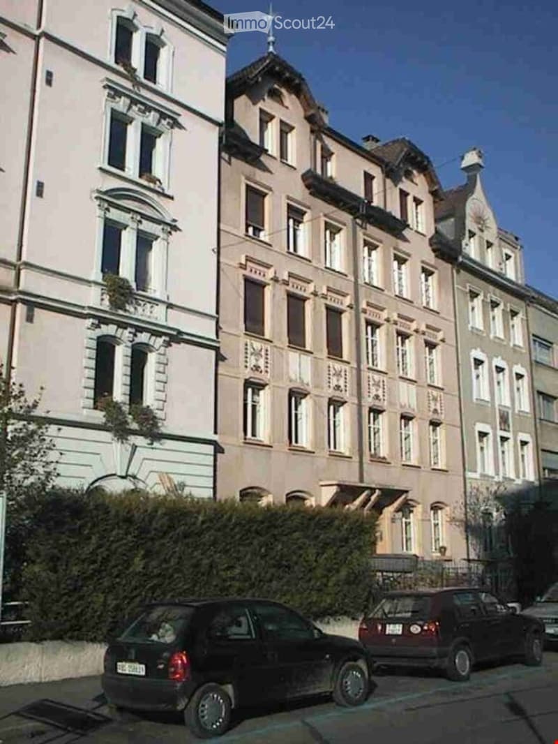 Multi-story residential building with classic European architecture, multiple windows, and balconies. Two cars are parked in front.
