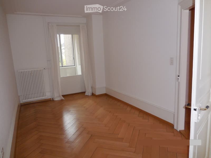 Empty room with wooden flooring, white walls, a window with curtains, and a radiator. No furniture or equipment is present.