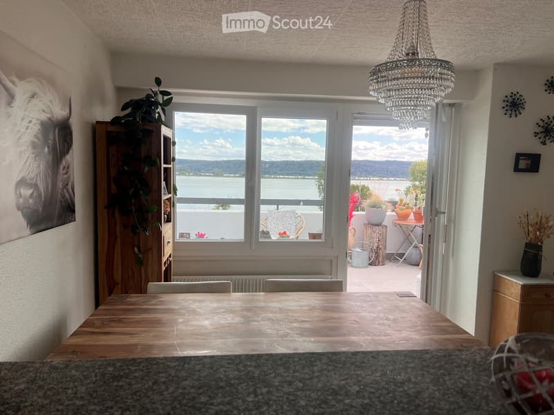 Dining area with a wooden table, chairs, cabinet, and a view of a lake through large windows and a sliding door leading to a balcony.