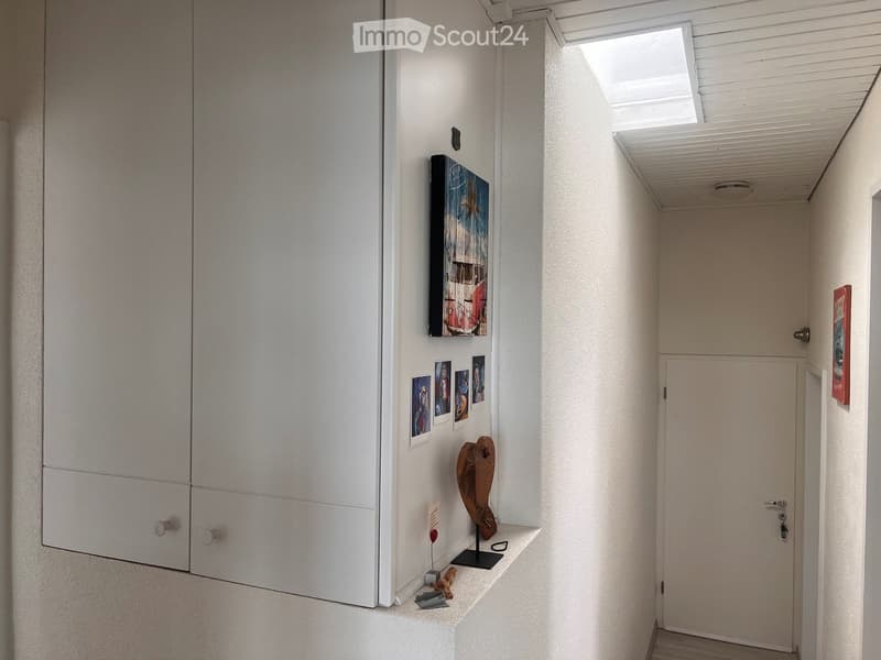 A narrow hallway with white walls, a built-in closet on the left, and a small shelf with decorative items and framed photos on the right. The ceiling has a skylight and recessed lighting.