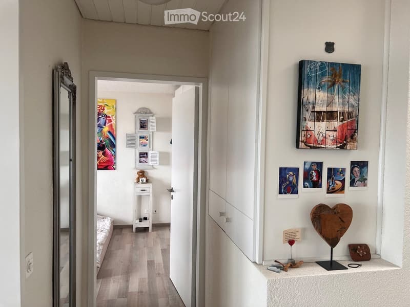 A narrow hallway with white walls, wooden flooring, and various decorative items including artwork, a mirror, and small shelves. The hallway leads to another room with a bed visible in the background.