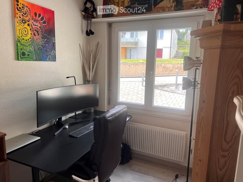 A well-lit room with a desk setup, a large monitor, laptop, keyboard, and a comfortable chair. A colorful painting hangs on the wall, and a vase with dried plants is placed on the shelf. A floor lamp is positioned near the window, and a radiator is visibl