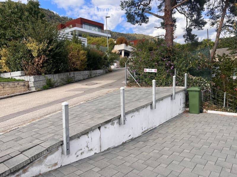 A paved outdoor area with a low white retaining wall, metal fence, and green trash bin. The area is bordered by a sidewalk and lush greenery, with modern houses visible in the background.