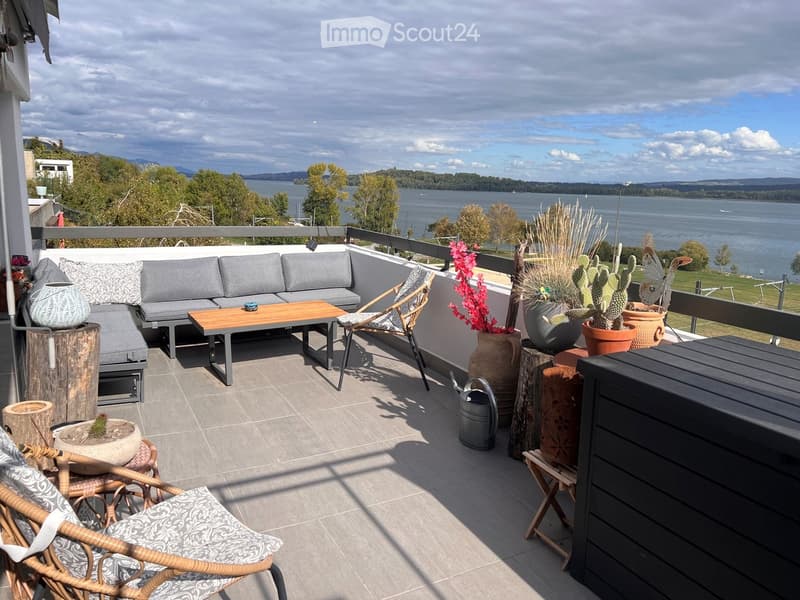 Balcony with a view of a lake, featuring a grey sofa, wooden coffee table, wicker chairs, and various potted plants.