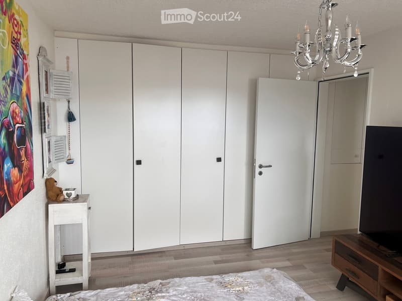Modern bedroom with a large white wardrobe, a bed with a decorative blanket, a small white table with a teddy bear, a TV stand, and a chandelier.