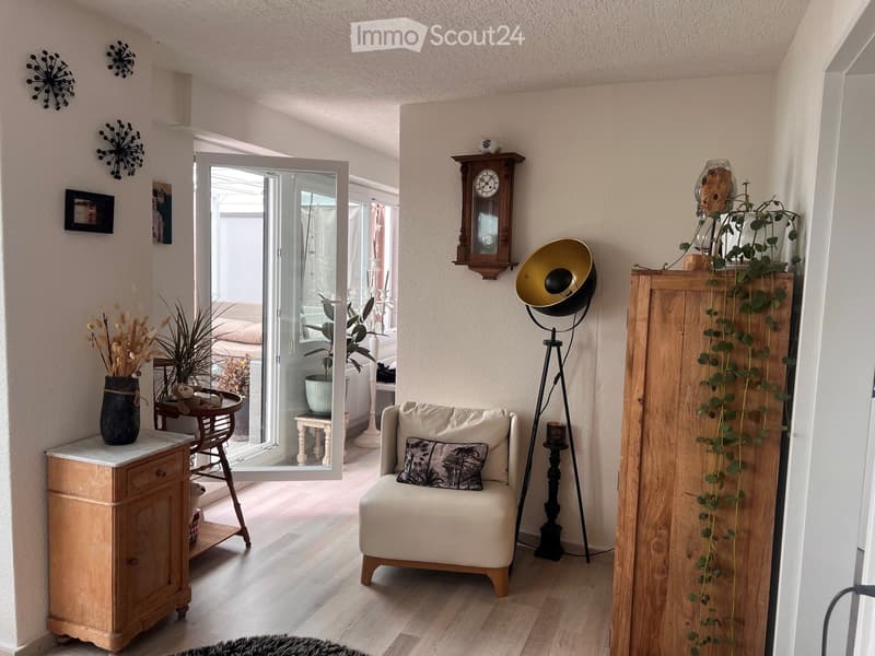 A cozy living space with a white armchair, a small wooden table with a vase of dried flowers, a floor lamp, a wooden cabinet with a tall plant, and a wall-mounted clock. The room features light wooden flooring and has an open doorway leading to another ro
