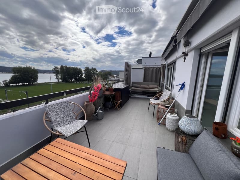 Balcony with a sofa, chairs, a wooden table, flower pots, and vases, overlooking a scenic view of a lake and greenery.
