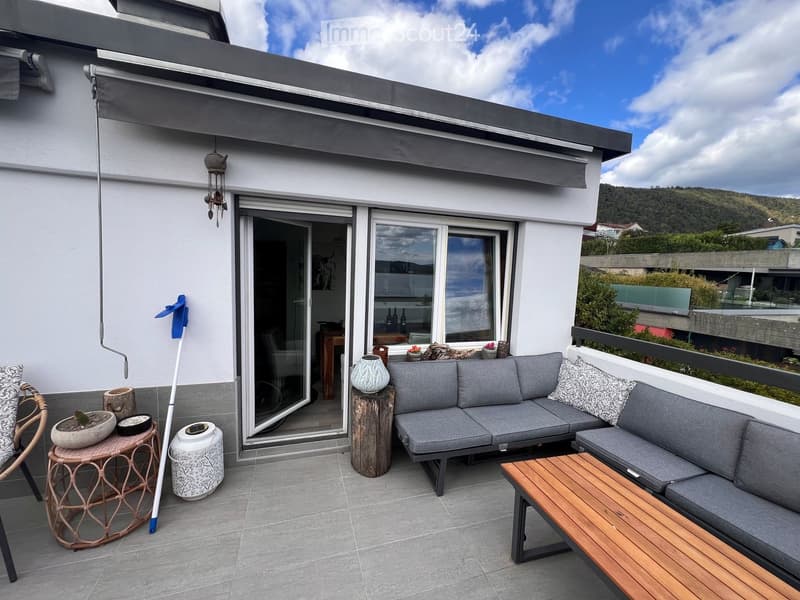 Balcony with grey sectional sofa, wooden coffee table, wicker chair, decorative pots, and vases.