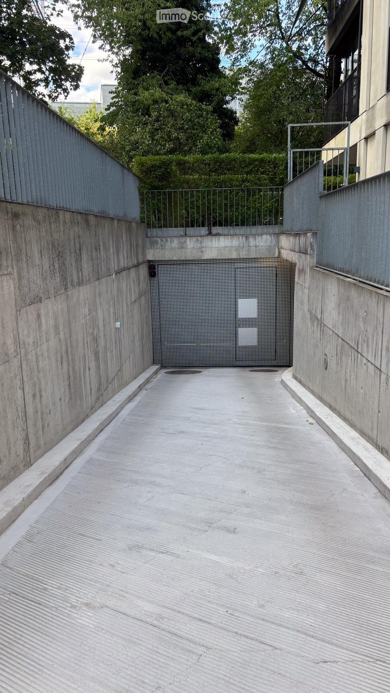 A narrow concrete corridor with metal railings on both sides, leading to a gated area with greenery visible beyond.