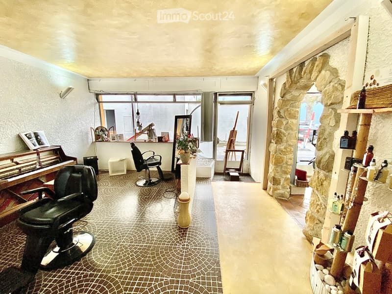 A well-lit salon with two black barber chairs, a small table with a plant, shelves with bottles, and a mirror. The floor has a decorative tile pattern, and there is a large window letting in natural light.