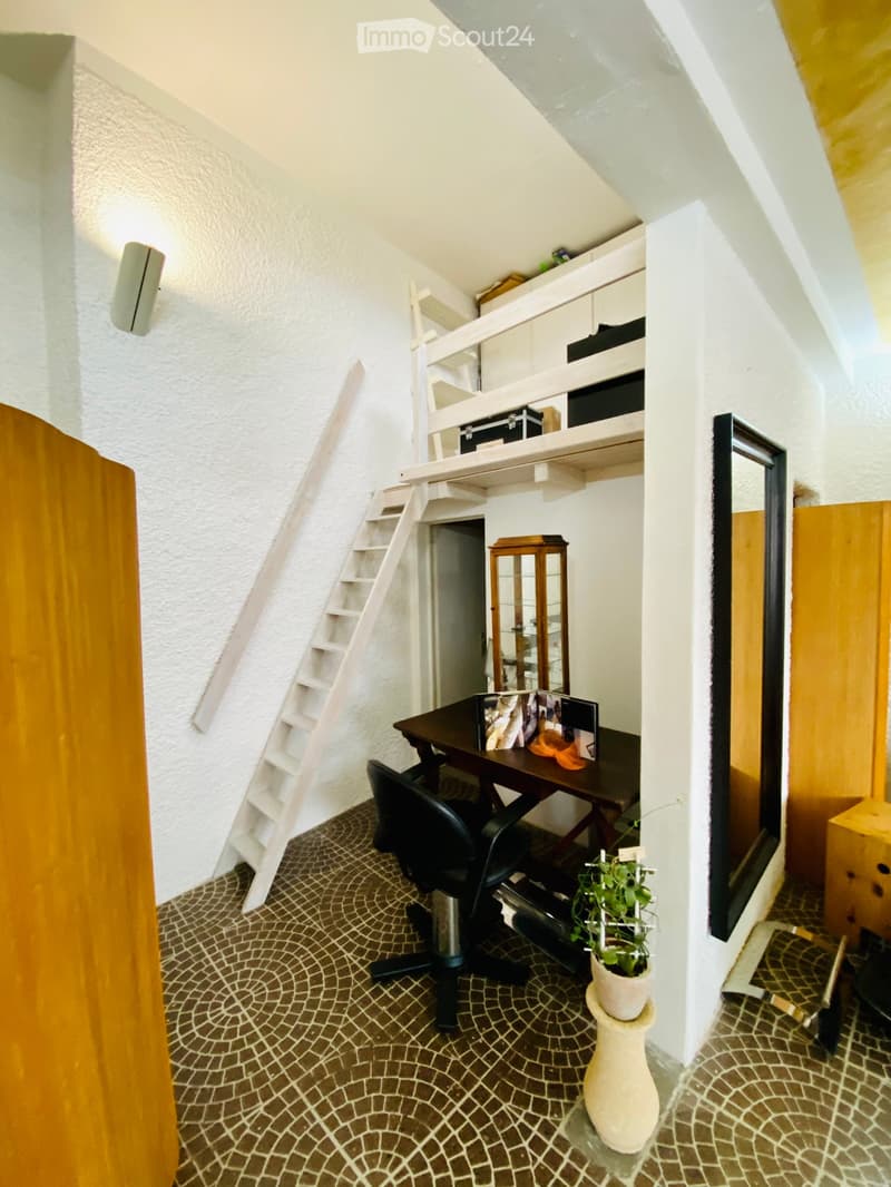 Cozy attic room with a slanted ceiling, featuring a wooden desk, black office chair, potted plant, and a small staircase leading to a lofted area with shelves and storage boxes.